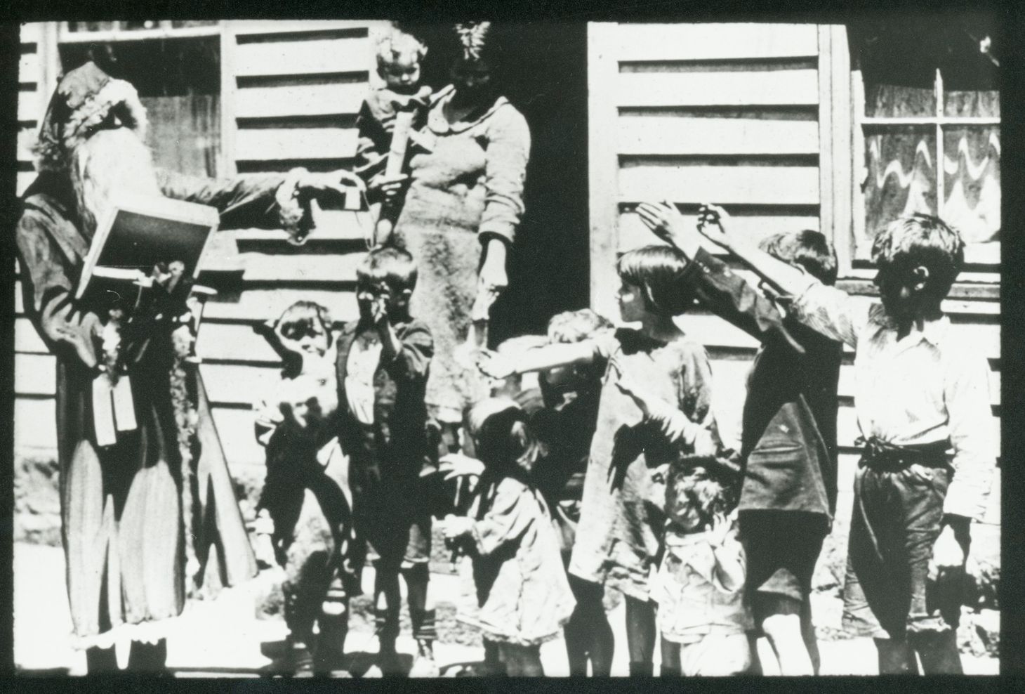 A black and white photo of Santa handing out presents to a bunch of kids outside an old, weatherboard house.