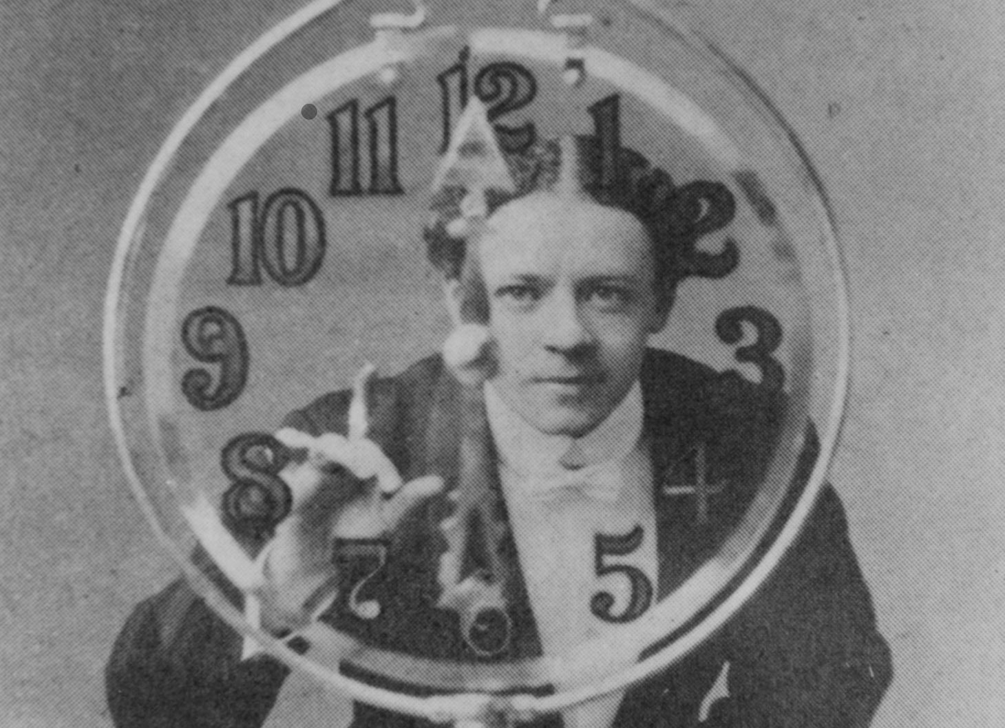 An honestly weird photo of a man with a severe middle-part hairstyle staring at you through a clear-glass clock.